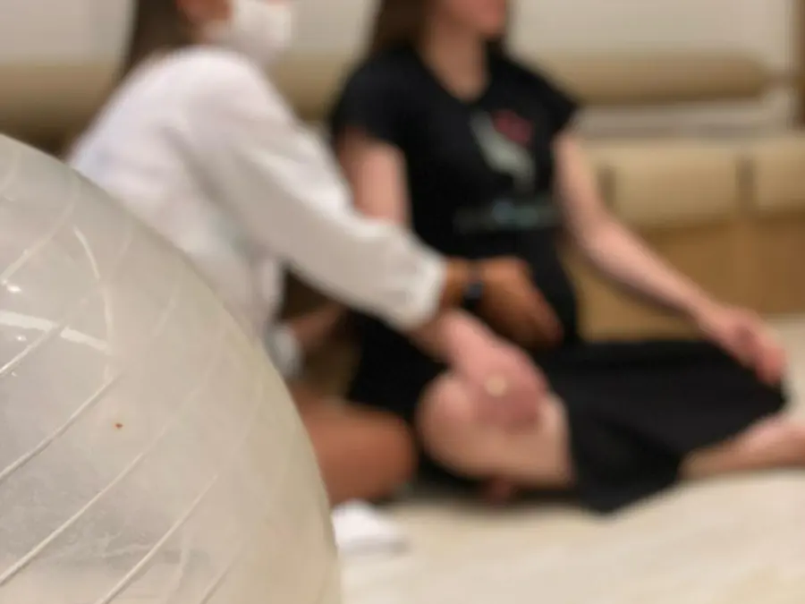 Fisioterapeuta de jaleco branco atende uma paciente sentada no tatame ao lado de uma bola de pilates, em foto desfocada que preserva a privacidade