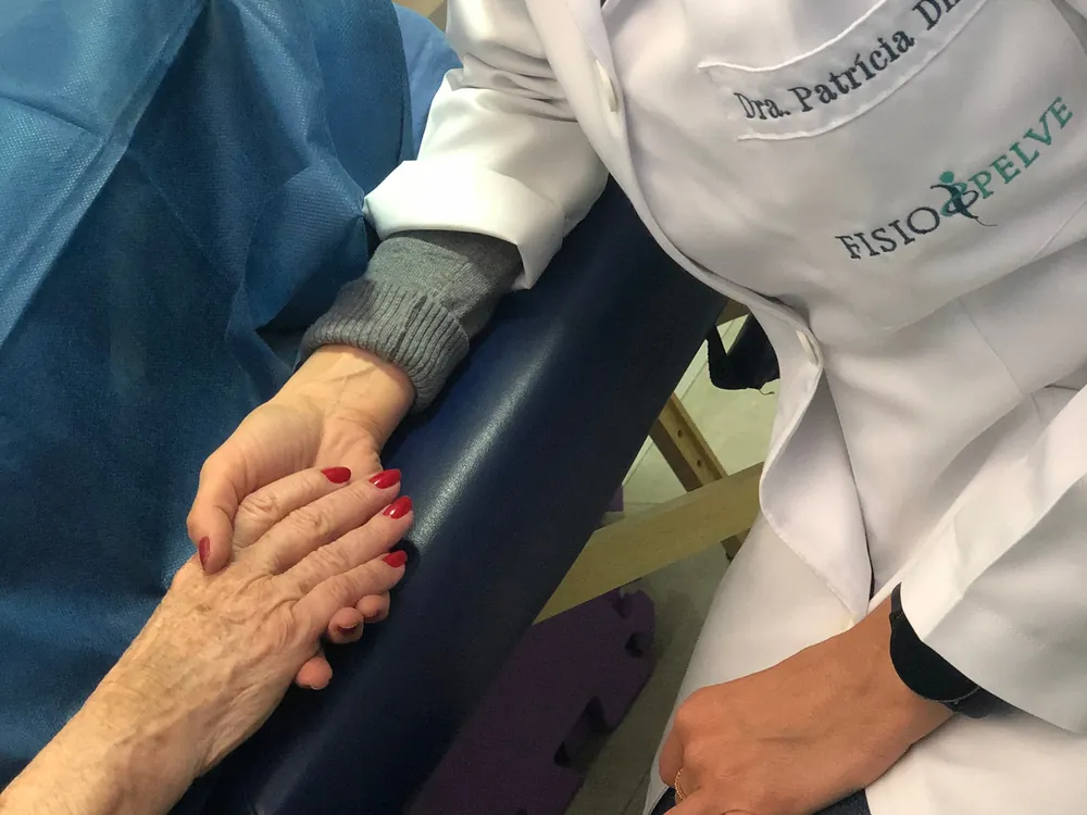 Dra. Patrícia Diniz, de jaleco branco com o bordado da Fisio&Pelve, segura a mão de uma paciente idosa durante o atendimento